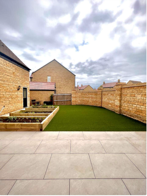 A landscaped backyard featuring artificial grass, wooden planter boxes, and a stone wall with neighboring houses in the background under a cloudy sky.